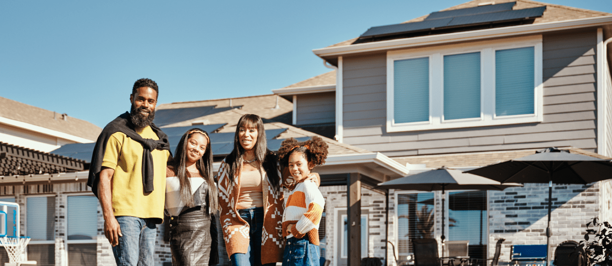A smiling family stands in front of their house with rooftop solar panels